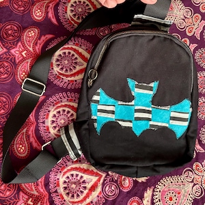 May include: Black crossbody bag with a bat applique in shades of blue, white, and black. The bag has a black adjustable strap and a zipper closure. The background is a purple paisley pattern.