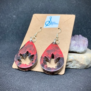 Può includere: Orecchini a goccia con un design a foglia d'acero rosso e nero. Gli orecchini hanno un bordo color argento e una foglia d'acero ritagliata al centro. Sono esposti su una scheda marrone con un logo.
