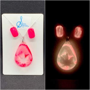 Puede incluir: Un conjunto de collar y pendientes de color rosa que brilla en la oscuridad con un colgante de hoja de arce. El colgante tiene forma de lágrima con una hoja de arce blanca en su interior. Los pendientes son pequeños y rectangulares.