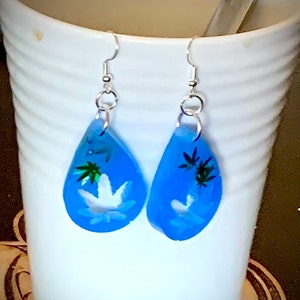 Peut inclure: Une paire de boucles d'oreilles en forme de larme bleue avec des crochets argentés. Les boucles d'oreilles présentent un motif de feuille de marijuana blanche avec des accents verts.