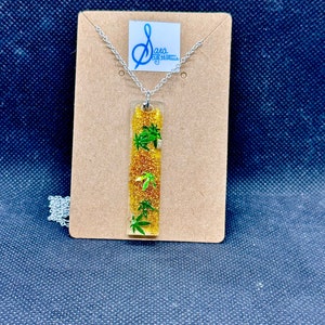 May include: A silver chain necklace with a gold and green rectangular pendant. The pendant is filled with gold glitter and green marijuana leaf shapes.