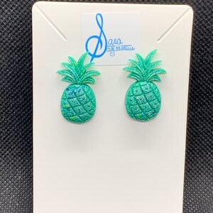 Op de afbeelding: Een paar groene glitter ananas oorbellen. De oorbellen zijn op een witte kaart met de tekst "Sara Sagittarius" erop.