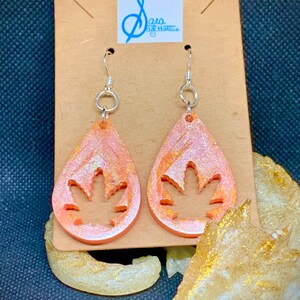 Könnte beinhalten: Ein Paar Hängeohrringe mit einer tropfenförmigen Form aus peachfarbenem Harz. Jeder Tropfen hat ein ausgeschnittenes Design eines Marihuana-Blattes. Die Ohrringe hängen an einer braunen Karte mit dem Text "Sara Sagittarius" darauf.