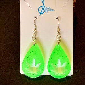 Puede incluir: Un par de pendientes verdes en forma de lágrima con un diseño de hoja de marihuana blanca. Los pendientes están en una tarjeta blanca con el texto "Sara Sagittarius" en azul.
