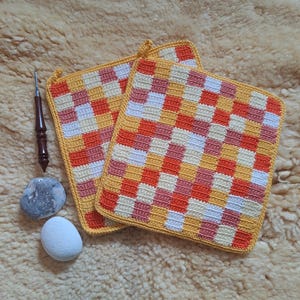 Könnte beinhalten: Zwei quadratische, handgefertigte Topflappen aus Häkelarbeit mit einem Schachbrettmuster in Orange-, Gelb-, Weiß- und Rosatönen. Ein Häkelhaken, ein grauer Stein und ein weißer Stein sind ebenfalls sichtbar. Die Topflappen haben einen gelben Rand.