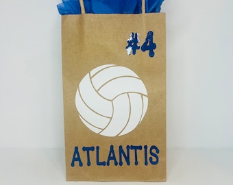May include: A brown paper gift bag with a white volleyball graphic and the text "#4 ATLANTIS" in blue. The bag is filled with blue tissue paper.