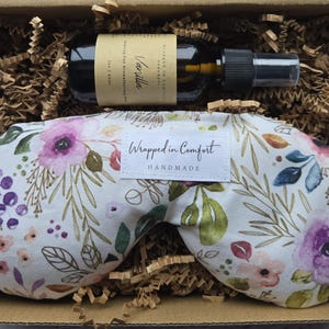 May include: A gift box with a floral eye mask and vanilla aromatherapy spray. The eye mask features a white background with a watercolour floral design in purple, pink, and green. The spray bottle is dark with a black sprayer. The box is filled with brown crinkle paper.