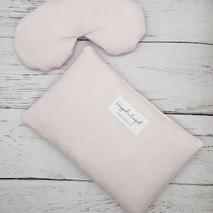 May include: A pink eye mask and a pink rectangular pillow with the text "Wrapped in Comfort - HANDMADE" on a white wooden surface.