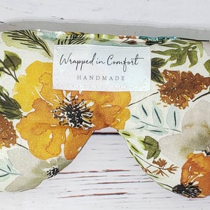 May include: A white eye mask with a floral pattern in shades of yellow, brown, and green. The eye mask has a white label that says "Wrapped in Comfort HANDMADE".