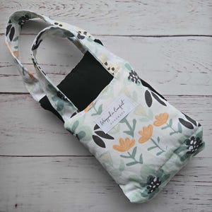 Könnte beinhalten: Eine Tragetasche mit floralem Muster, schwarzem Innenraum und Griff. Die Tasche hat einen weißen Hintergrund mit einem Muster aus grünen, schwarzen und orangefarbenen Blumen. Der Text "Wrapped in Comfort Handmade" befindet sich auf einem weißen Etikett.