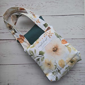 Könnte beinhalten: Eine Tragetasche mit floralem Muster und dunkelgrünem Innenraum. Die Tasche hat einen weißen Hintergrund mit großen, weichen gelben und orangefarbenen Blumen und grünen Blättern. Die Tasche hat eine rechteckige Form und einen Griff. Das Etikett lautet "Wrapped in Comfort, Handmade."