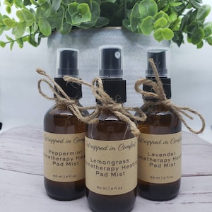 May include: Three brown glass bottles with spray tops and brown paper labels. The labels have the text "Wrapped in Comfort" and list different aromatherapy scents: Peppermint, Lemongrass, and Lavender. Each bottle contains 60 ml or 2 fl oz of liquid.