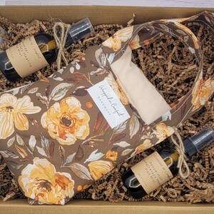 Peut inclure: Une boîte-cadeau remplie de papier de soie brun, un petit sac fourre-tout brun à motif floral et deux petites bouteilles d'huile essentielle. Le sac fourre-tout porte une étiquette indiquant "Wrapped in Comfort Handmade". Les bouteilles sont étiquetées "Lavender Happy Heating Body Mist" et "Peppermint Aromatherapy Pad Mist".