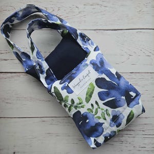 Könnte beinhalten: Eine Weinflaschentasche aus Stoff mit blau-grünem Blumenmuster auf weißem Grund. Sie hat ein marineblaues Innenfutter und einen Henkel. Das Etikett lautet "Wrapped in Comfort Handmade".