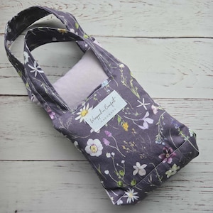 Könnte beinhalten: Eine kleine, rechteckige Tragetasche mit einem Blumenmuster in Lila, Weiß und Gelb. Die Tasche hat einen hellvioletten Innenraum und zwei Griffe. Ein weißes Etikett lautet "Wrapped in Comfort Handmade".