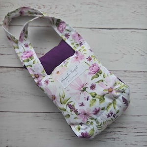 Könnte beinhalten: Eine Tragetasche mit floralem Muster und lila Innenraum. Die Tasche hat einen weißen Hintergrund mit rosa und lila Blumen und grünen Blättern. Der Griff besteht aus dem gleichen floralen Stoff. Die Tasche hat ein Etikett mit der Aufschrift "Wrapped in Comfort Handmade."