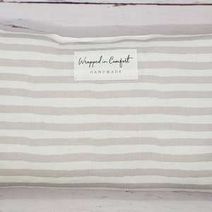 Peut inclure: Coussin rectangulaire à rayures horizontales grises claires et blanches. L'étiquette indique "Wrapped in Comfort HANDMADE". Il s'agit probablement d'un article de décoration intérieure.