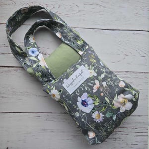 Könnte beinhalten: Graue Tragetasche mit floralem Muster, in der sich ein grüner Gegenstand befindet. Die Tasche hat einen Henkel und ein Etikett mit der Aufschrift "Wrapped in Comfort Handmade". Das florale Design umfasst weiße, blaue und rosa Blumen.