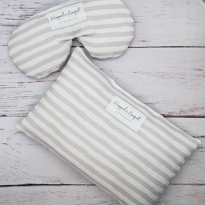 May include: Two striped fabric items, a contoured eye mask and a rectangular pillow, are displayed on a light wood surface. Both items feature a gray and white striped pattern and a small white label that reads "Wrapped in Comfort HANDMADE".