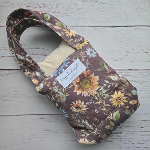 Könnte beinhalten: Eine Weinflaschentasche mit Blumenmuster und Henkel. Die Tasche besteht aus braunem Stoff mit einem Muster aus Sonnenblumen, Gänseblümchen und anderen Blumen in Gelb, Blau und Grün. Das Etikett lautet "Wrapped in Comfort Handmade."
