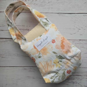 Könnte beinhalten: Eine Tragetasche mit floralem Muster und Griff. Die Tasche hat einen weißen Hintergrund mit orangefarbenen, gelben und grünen Blumenmustern. Auf der Vorderseite ist ein weißes Etikett mit dem Text "Wrapped in Comfort Handmade" aufgenäht.