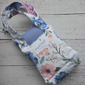 Könnte beinhalten: Flaschenträger mit floralem Muster und Griff. Der Stoff zeigt blaue, rosa und lila Blumen auf weißem Grund. Der Träger hat ein hellblaues Innenfutter und ein Etikett mit der Aufschrift "Wrapped in Comfort Handmade."