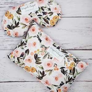 May include: A floral eye mask and a rectangular pillow, both featuring a watercolor design of pink roses, orange accents, and green leaves on a white background. The eye mask and pillow have a label that reads "Wrapped in Comfort HANDMADE".