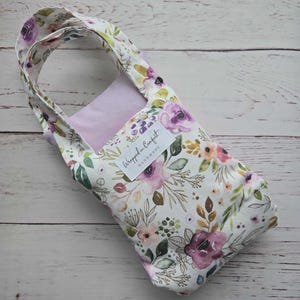 Könnte beinhalten: Tragetasche mit floralem Muster und lavendelfarbenem Innenraum. Die Tasche hat einen weißen Hintergrund mit lila, rosa und gelben Blumen und grünen Blättern. Sie hat einen Griff und ein Etikett mit der Aufschrift "Wrapped in Comfort Handmade."