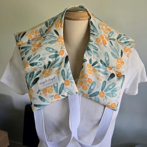 Peut inclure: Un coussin chauffant pour le cou et les épaules en tissu crème avec un motif aquarelle de baies orange et de feuilles bleu-vert. Il comporte deux sangles en tissu blanc et une étiquette blanche marquée Wrapped in Comfort HANDMADE.