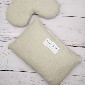 May include: Two beige linen eye pillows, one shaped like a crescent moon and the other rectangular. The rectangular pillow has a white label that reads "Wrapped in Comfort Handmade".