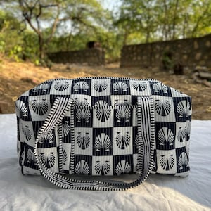 May include: A rectangular tote bag with a black and white seashell pattern. The bag has a striped handle and trim. The bag is sitting on a white surface, with a blurred background of trees and a stone wall.