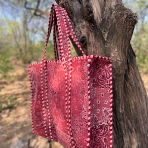 May include: A red tote bag with a paisley pattern and white trim. The bag has two handles and is hanging from a tree branch. The bag is approximately 38 cm wide and 30 cm tall.