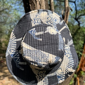May include: A black and white patterned bucket hat with a quilted design. The hat features a repeating pattern of geometric shapes and stylized figures in white and blue. The hat is hanging on a tree branch.