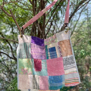Peut inclure: Sac à bandoulière patchwork avec une sangle rose. Le sac est composé de carrés de tissu de différentes couleurs : rose, violet, vert et bleu. Le sac est suspendu à une branche d'arbre.