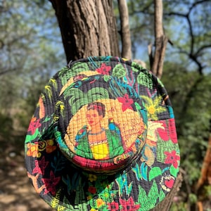 Op de afbeelding: Een gewatteerde zwarte zonnehoed met een levendig bloemen- en bladerenpatroon in rood, groen, geel en oranje. De hoed heeft een centraal beeld van een vrouw met papegaaien. De hoed hangt aan een tak.