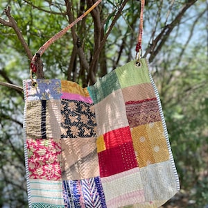 Puede incluir: Un bolso tote patchwork con un diseño acolchado colorido. El bolso presenta una variedad de cuadrados de tela en tonos de rojo, amarillo, verde y azul, con una correa estampada. El bolso está colgado de una rama de árbol.