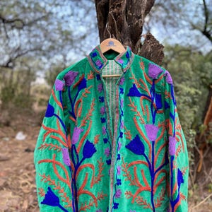May include: A green velvet jacket with floral embroidery in shades of purple, blue, and orange. The jacket is hanging on a wooden hanger against a blurred outdoor background. The embroidery creates a striking contrast against the jacket's vibrant green color.