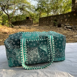 Könnte beinhalten: Gesteppte Reisetasche mit türkis-weißem Paisley-Muster und grün-weiß gestreiften Griffen. Die Tasche ist rechteckig und hat einen Reißverschluss oben. Sie liegt auf einer weißen Oberfläche.