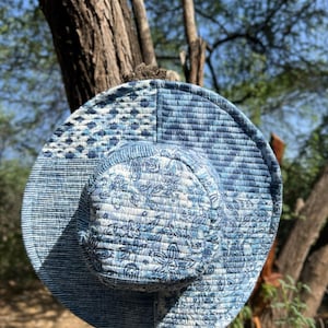 Puede incluir: Un sombrero para el sol azul estampado con ala ancha y diseño floral en la copa. El sombrero está hecho de tela acolchada y cuelga de una rama de árbol. La paleta de colores del sombrero incluye tonos de azul y blanco.