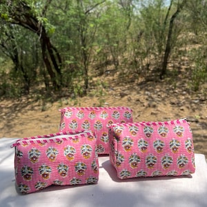 Op de afbeelding: Drie roze make-up tassen met een bloemenpatroon. De tassen zijn gerangschikt op een wit oppervlak, met een natuurlijke achtergrond van bomen en bladeren. Elke tas heeft een ritssluiting en een contrasterende rand.