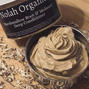 May include: A brown jar of Nolah Organics Marshmallow Root & Molasses Deep Conditioner. The jar is open and the product is in a silver bowl. The product is a thick, creamy consistency.