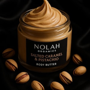 May include: A jar of Nolah Organics Salted Caramel & Pistachio Body Butter. The jar is filled with a swirl of light brown body butter and surrounded by several pistachios. The jar has a black label with white and brown text. The background is black.