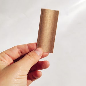 Op de afbeelding: Een klein, rechthoekig stuk lichtbruin hout dat in een hand wordt gehouden. De houtnerf is zichtbaar en loopt verticaal. Het hout lijkt een dun fineer of een klein stukje sierlijst te zijn. De achtergrond is effen wit.