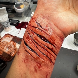 Puede incluir: Primer plano de una muñeca con múltiples cortes profundos y sangrientos. Los cortes son rojos y parecen frescos, con sangre visible. La piel alrededor de los cortes también está manchada de sangre. La imagen sugiere una aplicación de maquillaje de efectos especiales.