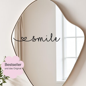 Könnte beinhalten: Ein unregelmäßig geformter Spiegel mit goldenem Rahmen. Das Wort "smile" ist in schwarzer Schrift über die Oberfläche des Spiegels geschrieben, wobei ein Herz den Buchstaben "i" ersetzt. Ein rosa blütenförmiger Aufkleber mit dem Text "Bestseller" und "und das Original" befindet sich in der unteren linken Ecke.