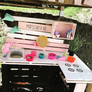 Könnte beinhalten: Eine rosafarbene und weiße Holzspielküche mit dem Schriftzug "Matschküche" auf der Vorderseite. Die Küche hat ein Spülbecken, einen Herd und ein Regal mit verschiedenen Spiellebensmitteln und -utensilien.