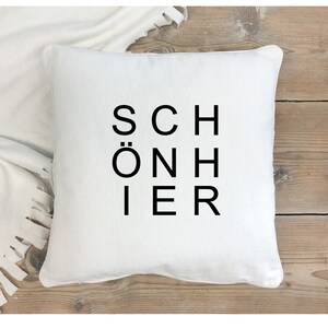Könnte beinhalten: Ein quadratisches weißes Kissen mit dem Wort "SCHÖNIER" in Schwarz vertikal gedruckt. Das Kissen liegt auf einer Holzoberfläche, mit einer weißen Decke im Hintergrund.