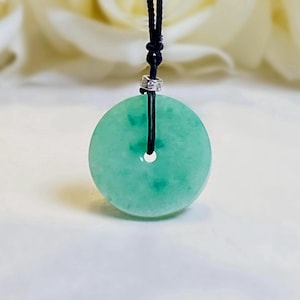 May include: A circular, light green jade-like stone pendant with a central hole, suspended from a thin black cord. A small, silver-toned metal bead secures the cord at the top of the stone, creating a simple, adjustable necklace design.