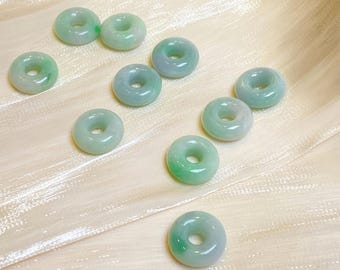May include: A collection of jade donut-shaped beads in varying shades of green. The beads are arranged on a cream-colored, textured surface. The beads have a smooth, polished finish and a hole in the center.