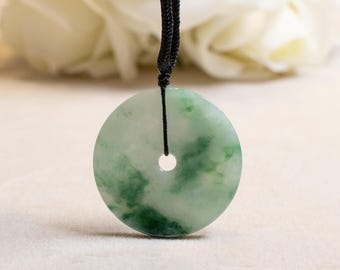 May include: A circular jade pendant, with a central aperture, hanging from a black cord. The pendant displays a blend of pale and deep green tones, producing a marbled appearance. The backdrop is blurred, featuring white flowers.